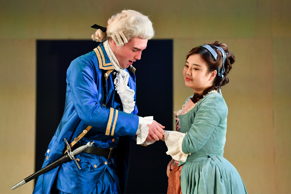 A male student, dressed in an elaborate blue costume, holding and singing to a female student, wearing a peasant costume with ribbons in her hair, in an opera performance.